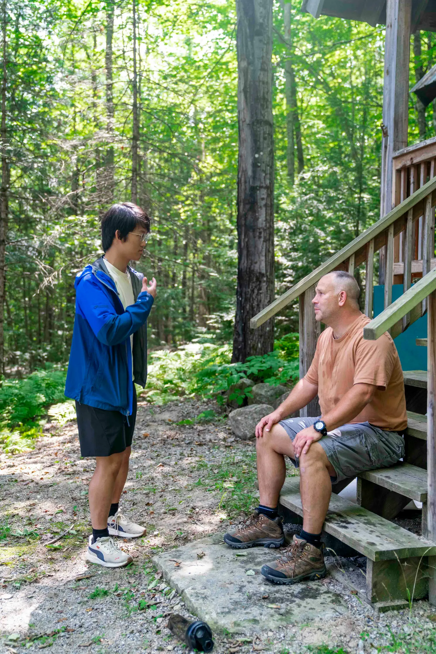 Person standing and speaking with another person seated on cabin steps in a wooded area"