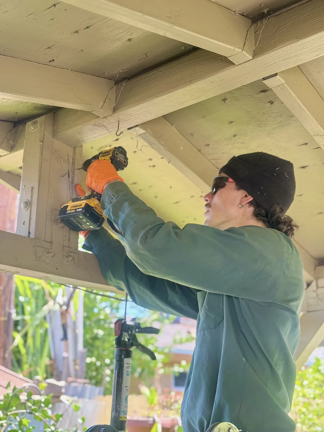 Treebark technician drilling patio beams during termite treatment, orange gloves and company patches